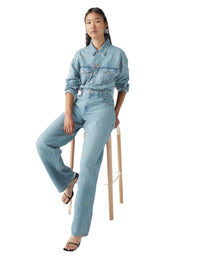 Person wearing a light blue denim jumpsuit sitting on a stool against a white background.