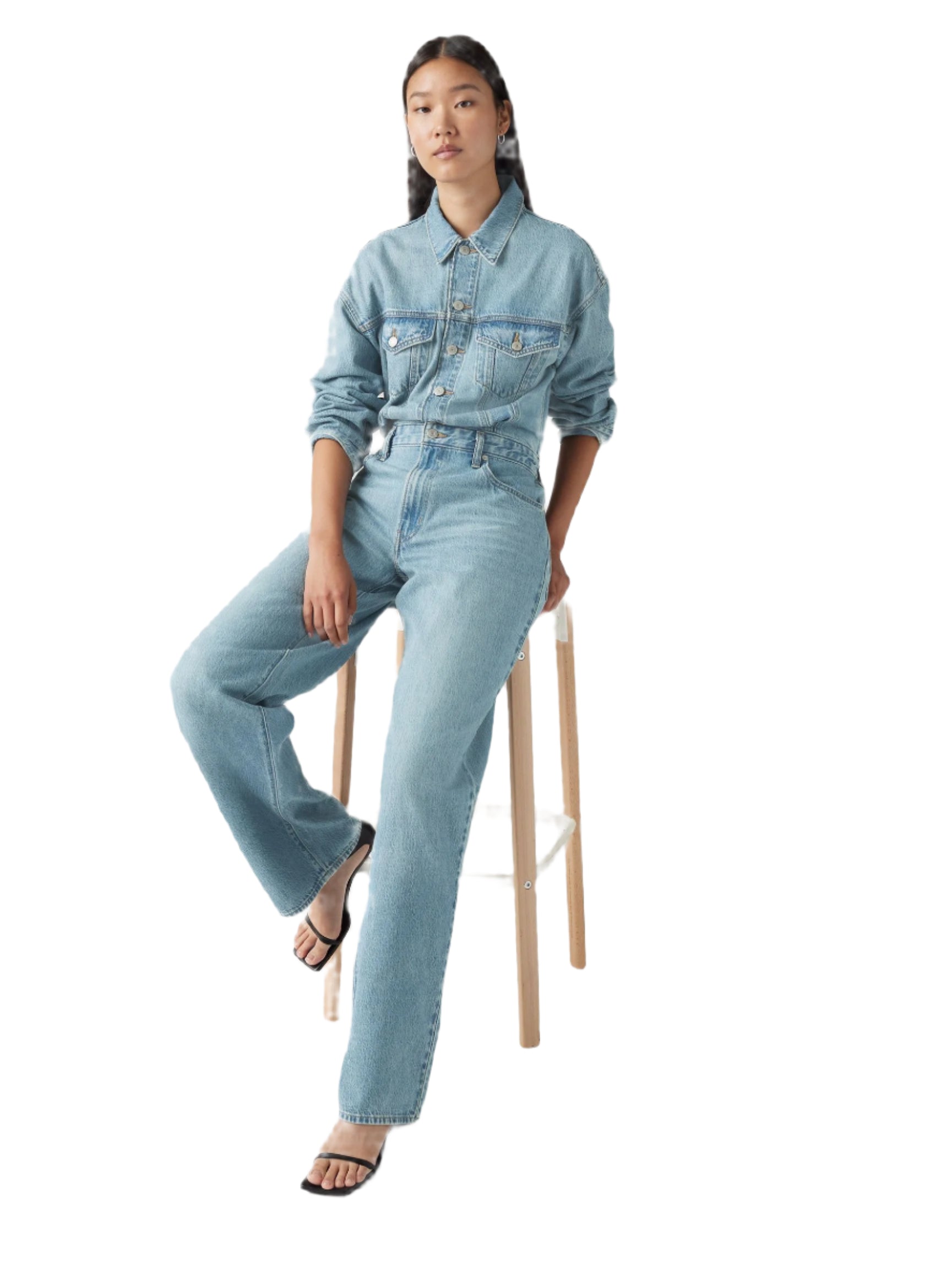 Person wearing a light blue denim jumpsuit sitting on a stool against a white background.