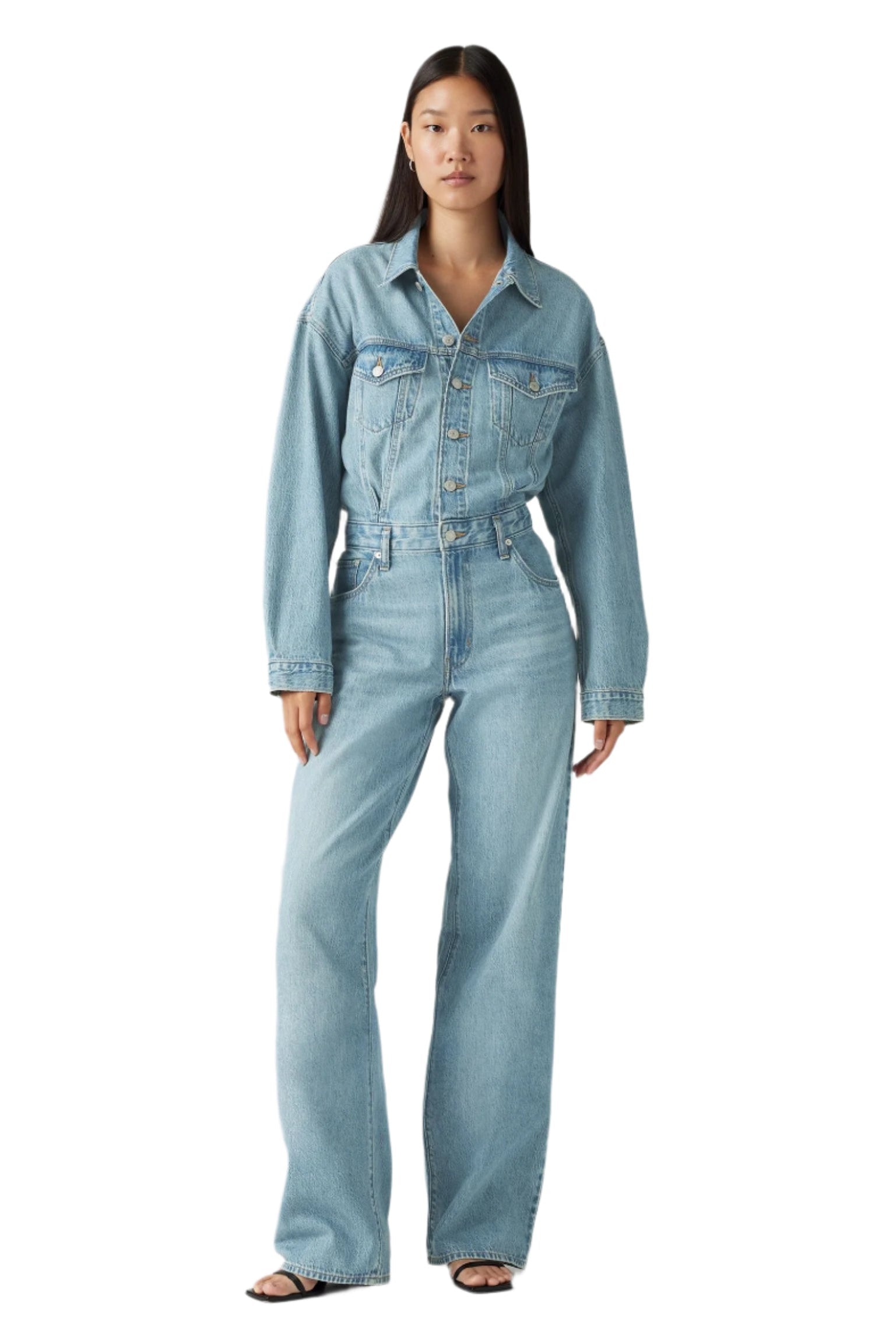 A woman standing against a white background, wearing a blue denim baggy jumpsuit with a collared neckline, long sleeves, and straight-leg design. She has a belt loops and the jumpsuit has chest, side, and back pockets.