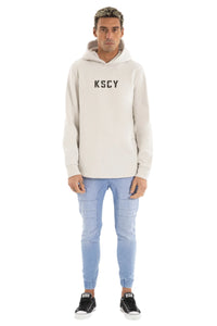 Man wearing a beige hoodie with 'KSCY' branding and light blue jeans on a white background