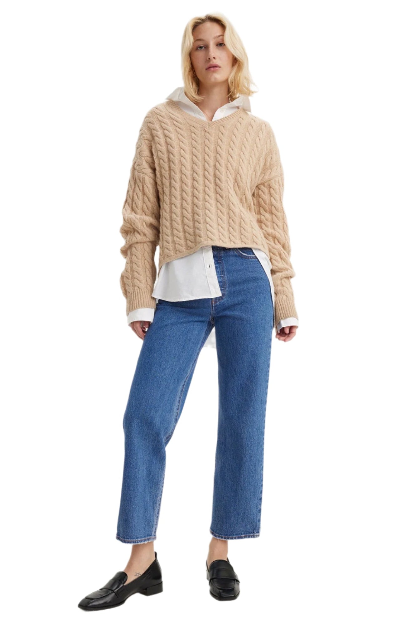 A woman is standing, wearing straight leg blue jeans and a beige cable knit sweater with a hoodie.