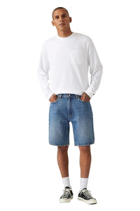 A man modeling a white long-sleeve crewneck shirt with a chest pocket, paired with blue denim shorts, white socks, and black-and-white sneakers, against a light gray studio background.