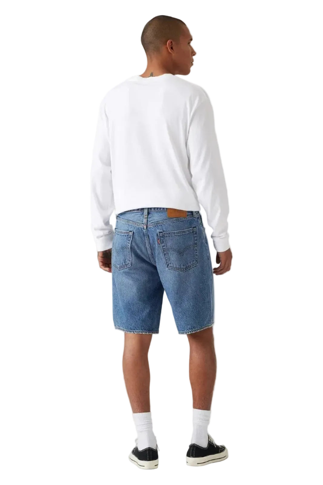 A man is shown from behind wearing a white long-sleeve crewneck sweatshirt, medium wash blue denim shorts, white socks, and black sneakers, photographed against a neutral light gray studio background.