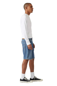 A man in profile wearing a white long-sleeve crewneck shirt, blue denim shorts, white socks, and black low-top sneakers, standing against a light gray studio background.