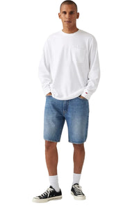 A man modeling a white long-sleeve crewneck pocket shirt, blue denim shorts, white socks, and black low-top sneakers against a light gray studio background.