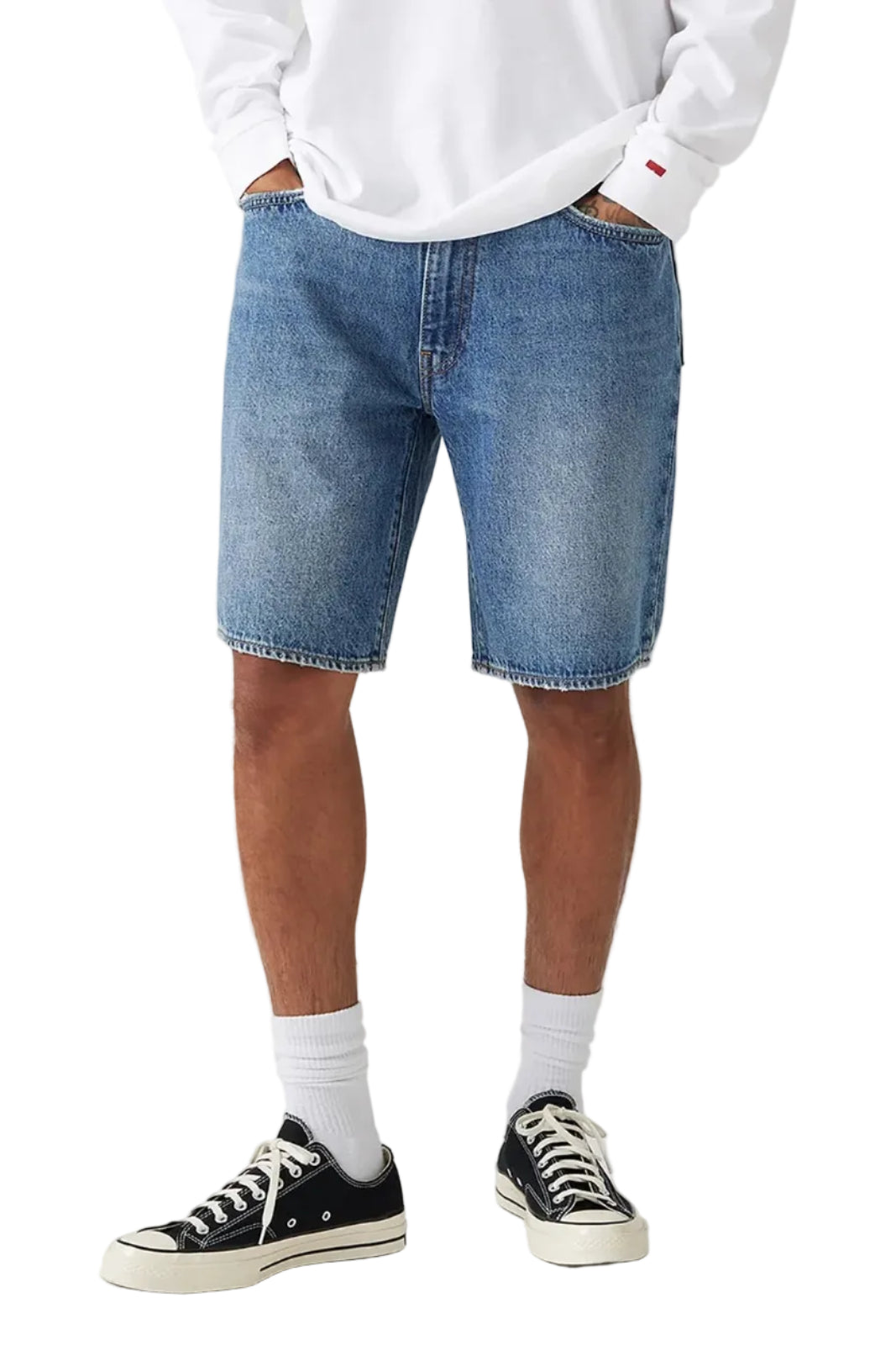 A person wearing light blue denim shorts, a white sweatshirt, white socks, and black low-top sneakers, standing against a light gray studio background.