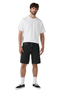 A man modeling a white crewneck t-shirt, black denim shorts, white socks, and black low-top sneakers against a light gray studio background.