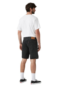 A man is shown from the back wearing a white t-shirt, black denim shorts, white socks, and black low-top sneakers against a light gray studio background.
