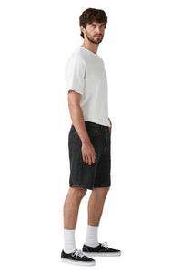 A man stands in profile wearing a plain white short-sleeve T-shirt, dark charcoal denim shorts, white socks, and black low-top sneakers against a light gray studio background.