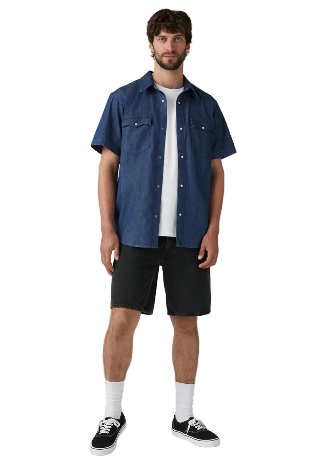 A man modeling a short-sleeve blue denim button-up shirt over a white t-shirt, paired with black shorts, white socks, and black sneakers, against a light gray studio background.