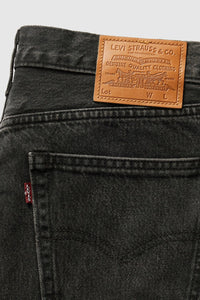 Close-up of the back waistband of Levi's 501 jeans in washed black denim, showing the signature brown leather patch with the brand name and a small red tab on the pocket.
