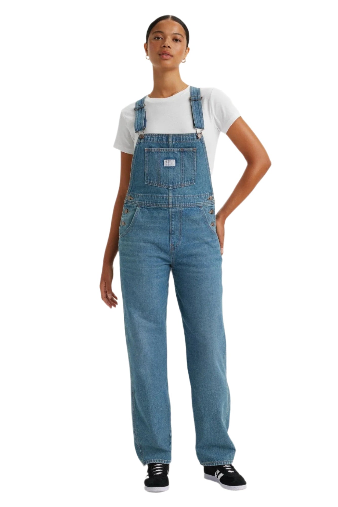 A woman standing, wearing vintage inspired blue denim overalls with adjustable straps, bib front with pockets, and straight leg cut.